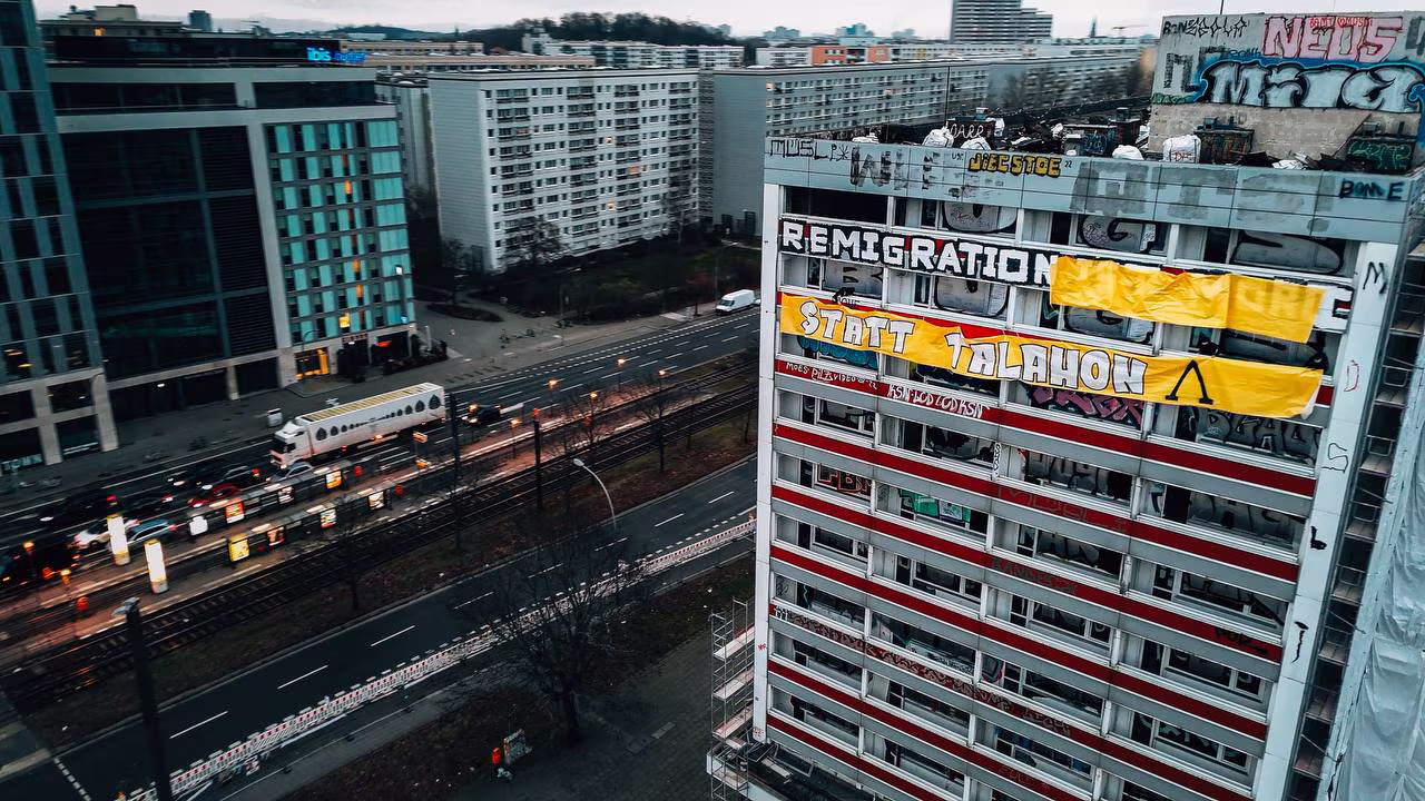Mehrstöckiges Wohngebäude in Berlin mit mehreren großen Bannern an der Fassade, darunter ein Banner mit der Aufschrift 'REMIGRATION STATT TALAHON', umgeben von Graffiti, im Hintergrund Straßen mit Verkehr und weiteren Wohngebäuden.